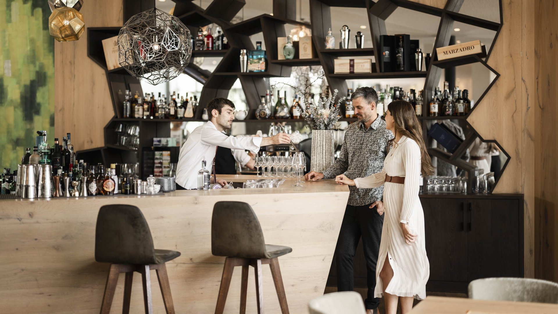 Schenna Resort Bar with bartender and two guests in front of a modern wooden shelf filled with bottles
